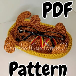 May include: A crocheted eyeglass holder in shades of brown and yellow, with the text "PDF Pattern" and "CrochetCustomsCA". The holder contains a pair of eyeglasses. The holder is designed to resemble a hot dog.