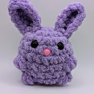 May include: A soft, purple crocheted bunny with black eyes and a pink nose. The bunny is made with a fluffy yarn and has a rounded body.