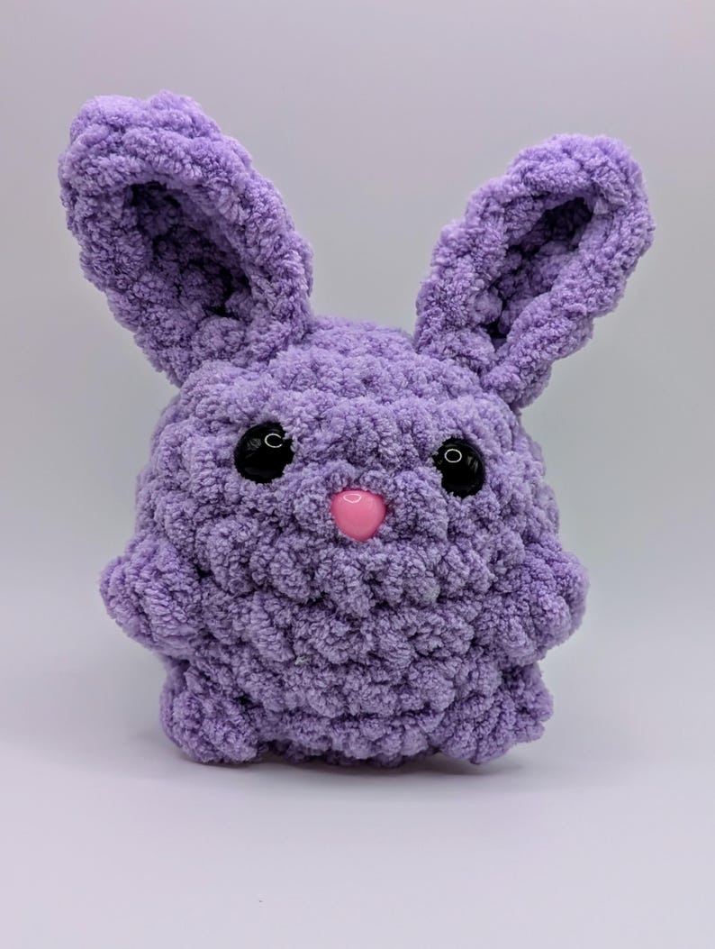 May include: A soft, purple crocheted bunny with black eyes and a pink nose. The bunny is made with a fluffy yarn and has a rounded body.