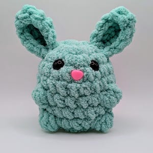 May include: A crocheted plush toy shaped like a bunny with large ears. The bunny is light blue with black eyes and a pink nose.
