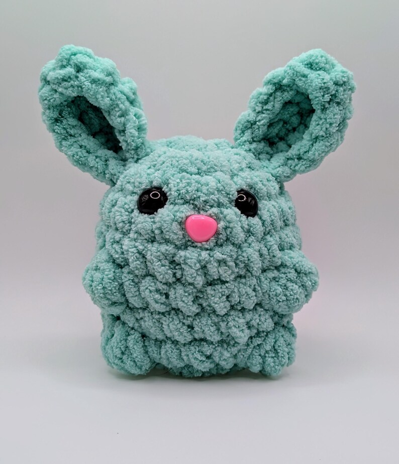 May include: A crocheted plush toy shaped like a bunny with large ears. The bunny is light blue with black eyes and a pink nose.