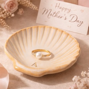 May include: A gold ring with a pearl center stone and small accent stones rests in a seashell-shaped dish. The dish is light yellow with a gold trim and the letter "A" at the base. A "Happy Mother's Day" card and a pink gift box are in the background.