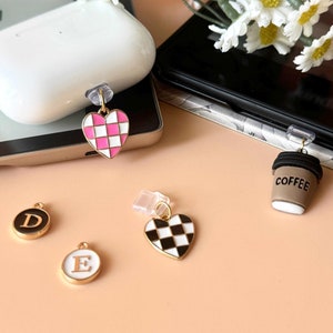 May include: Assortment of charms for phone cases, including a pink and white checkered heart, a black and white checkered heart, a coffee cup charm, and two letter charms, one with the letter D and one with the letter E.
