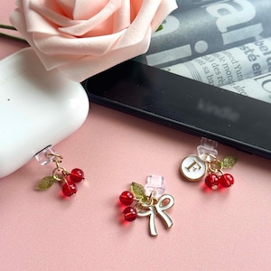 Puede incluir: Tres dijes decorativos con racimos de cuentas de cerezas rojas, hojas verdes y detalles dorados. Un dije presenta un lazo blanco, y otro tiene un círculo blanco con la letra "F". Los dijes están sujetos a clips transparentes.