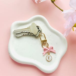 May include: A set of keys rests on a white, wavy-edged dish. A gold-colored keychain with a pink bow and a circular charm with the letter "E" hangs from the keys. The background is a soft pink with white flowers.