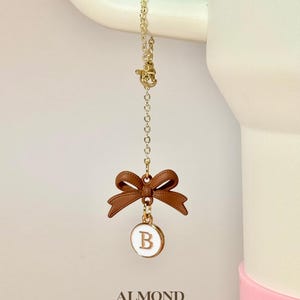 May include: A gold chain with a brown bow and a white circle charm with the letter "B" in gold. The charm is hanging from a white mug with the word "ALMOND" printed below it.