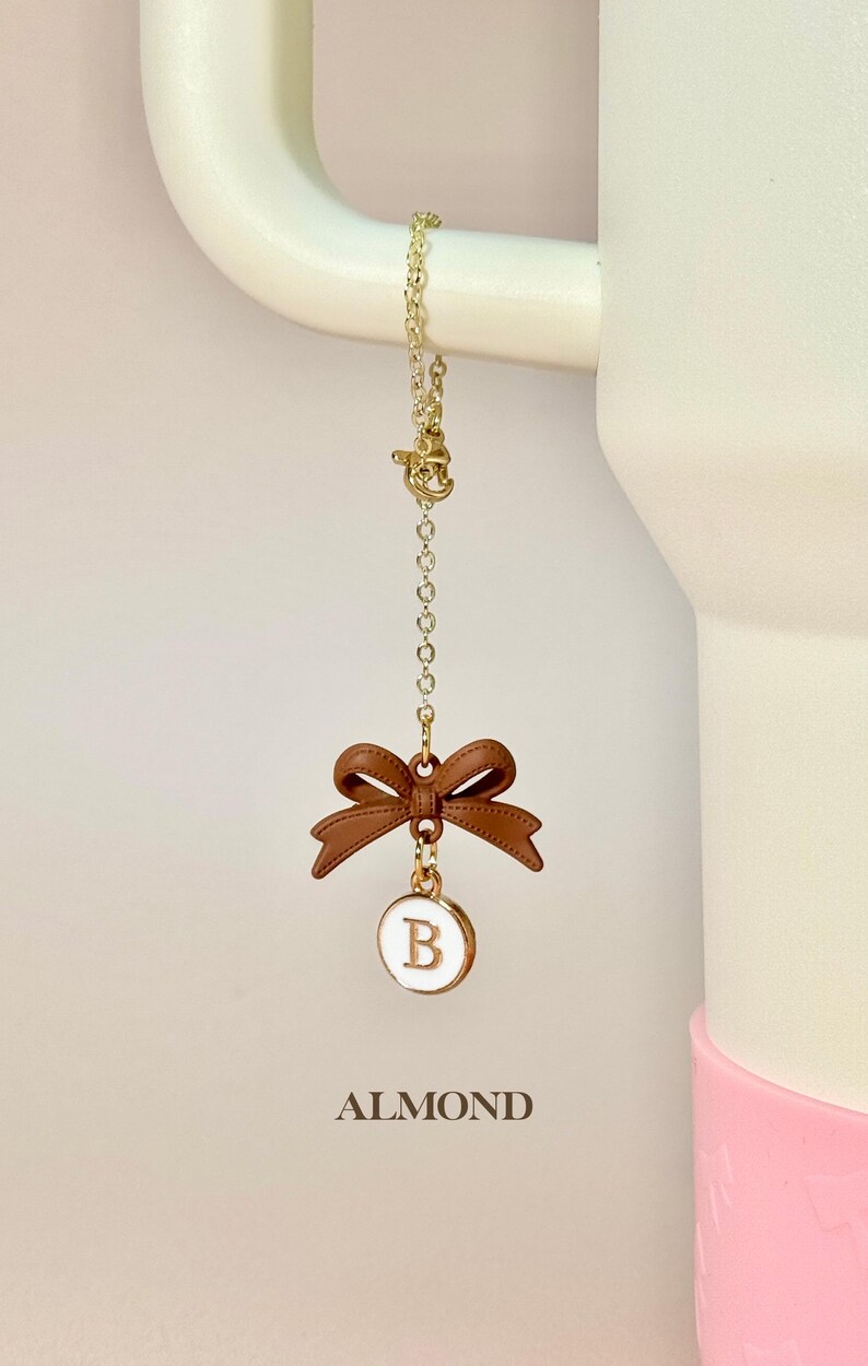 May include: A gold chain with a brown bow and a white circle charm with the letter "B" in gold. The charm is hanging from a white mug with the word "ALMOND" printed below it.