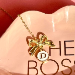 May include: A gold chain necklace with a gold bow charm and a white circle pendant with the letter "D" on a pink dish with the text "The Boss" in gold lettering.