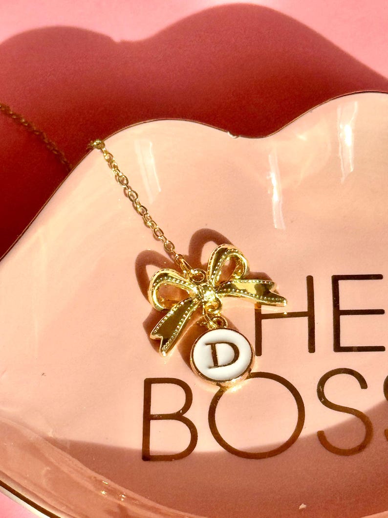 May include: A gold chain necklace with a gold bow charm and a white circle pendant with the letter "D" on a pink dish with the text "The Boss" in gold lettering.