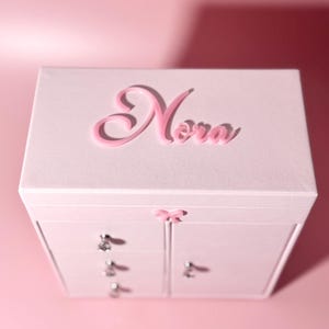 May include: A light pink jewelry box featuring the name Nora in raised pink script lettering on the top lid. The box includes three stacked drawers on the left side and a single door on the right, all accented with small, clear crystal knobs.