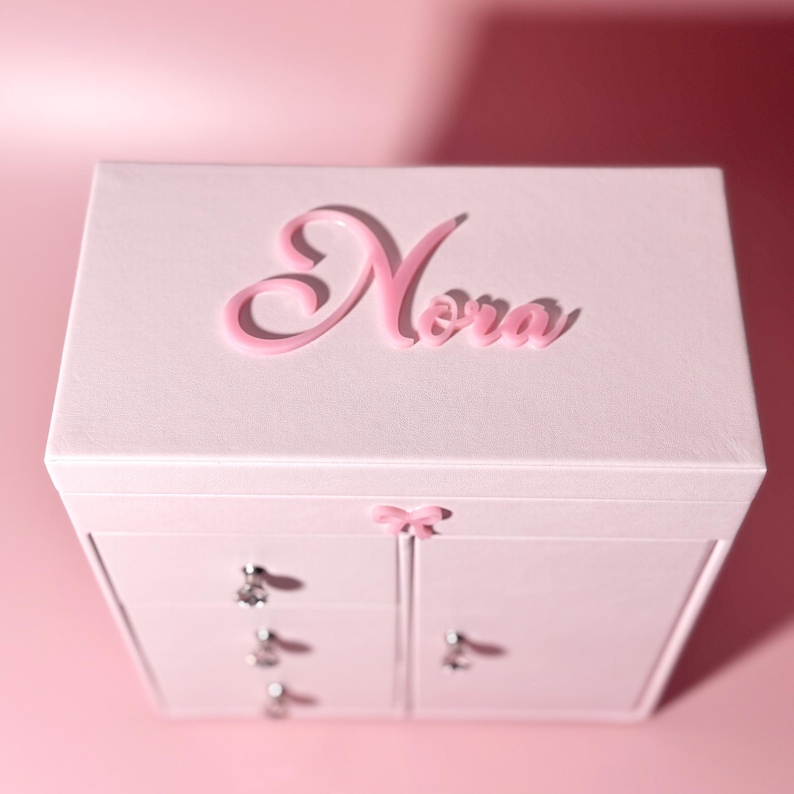 May include: A light pink jewelry box featuring the name Nora in raised pink script lettering on the top lid. The box includes three stacked drawers on the left side and a single door on the right, all accented with small, clear crystal knobs.