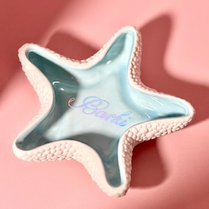 May include: A starfish-shaped ceramic trinket dish with a textured, bumpy white exterior and a smooth, glossy light blue interior. The center of the dish features the word Barki written in a cursive, light blue script font.