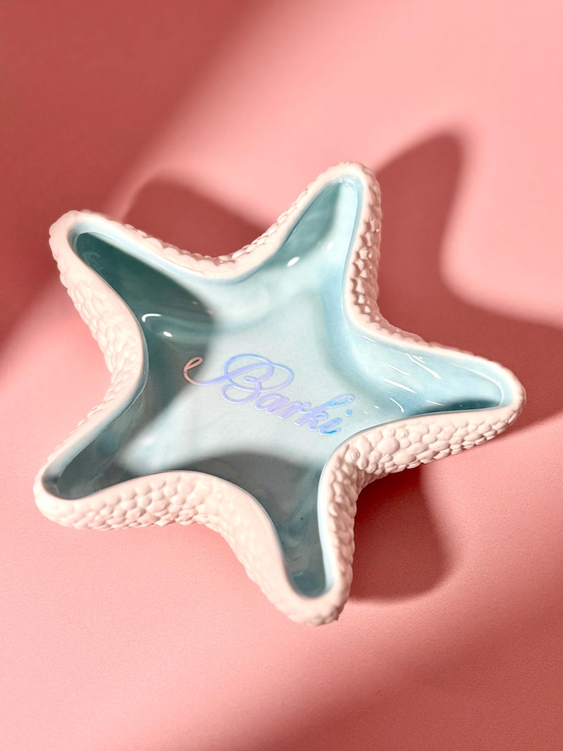 May include: A starfish-shaped ceramic trinket dish with a textured, bumpy white exterior and a smooth, glossy light blue interior. The center of the dish features the word Barki written in a cursive, light blue script font.