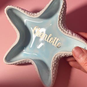 May include: A star-shaped ceramic trinket dish with a glossy light blue interior and a textured white exterior. The name Charlotte is written in gold script lettering across the center of the dish, which features five distinct points.