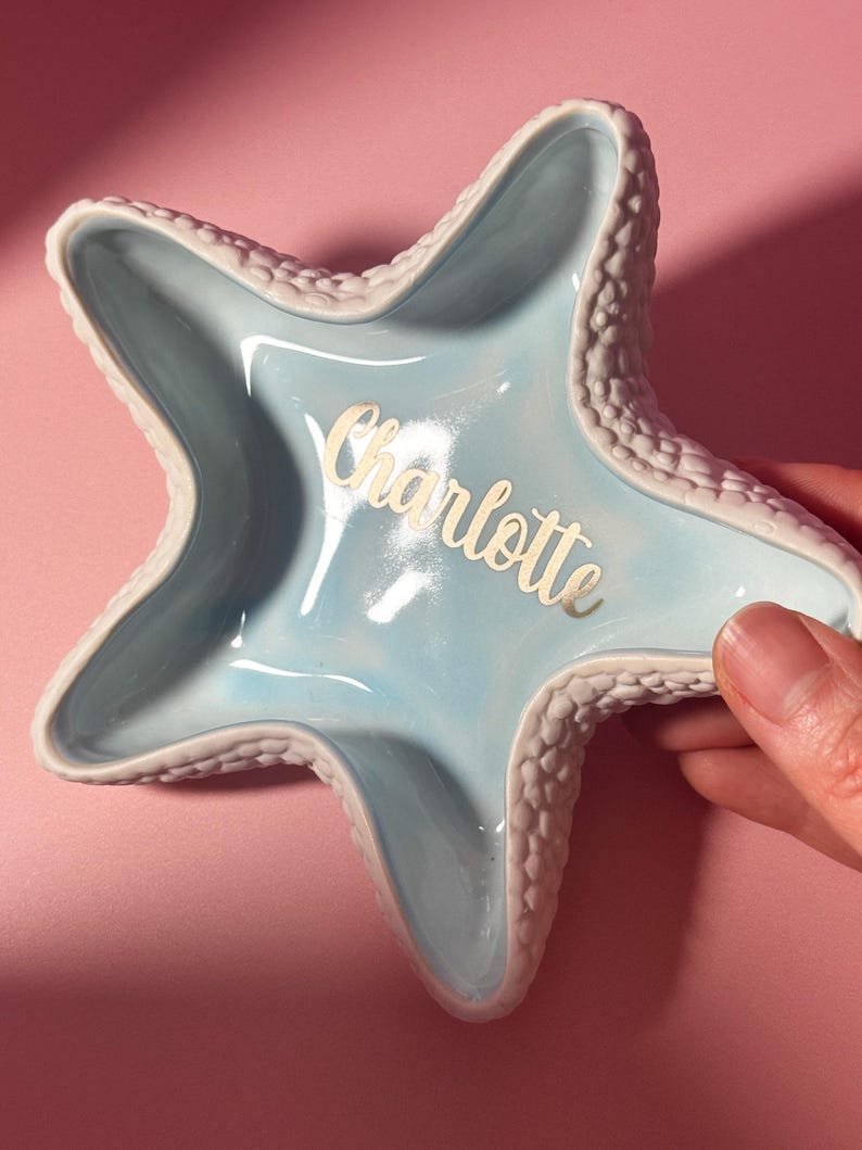 May include: A star-shaped ceramic trinket dish with a glossy light blue interior and a textured white exterior. The name Charlotte is written in gold script lettering across the center of the dish, which features five distinct points.