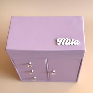 May include: A lilac jewelry box with drawers and a cabinet. The box has silver-coloured knobs and the name "Mila" in white script on the top. The box is on a light tan surface.