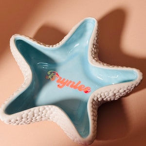 May include: A star-shaped ceramic trinket dish with a textured white exterior and a smooth, glossy light blue interior. The name Brynlee is written in the center in a holographic, iridescent script font that reflects multiple colors.