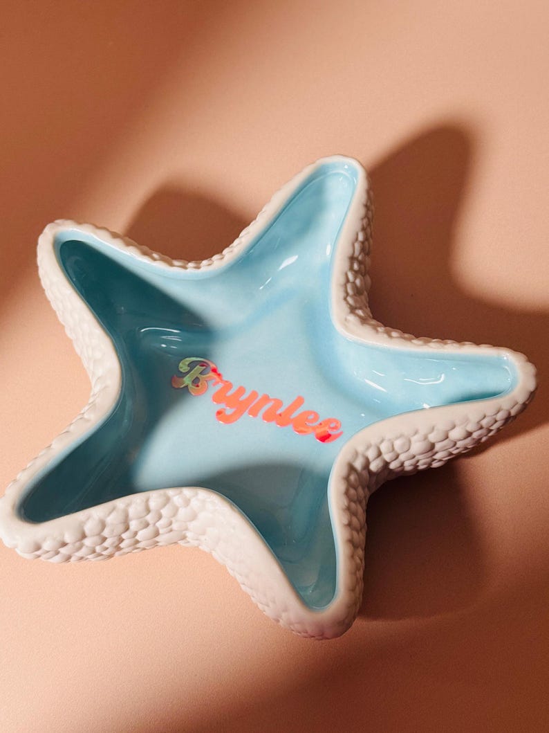 May include: A star-shaped ceramic trinket dish with a textured white exterior and a smooth, glossy light blue interior. The name Brynlee is written in the center in a holographic, iridescent script font that reflects multiple colors.