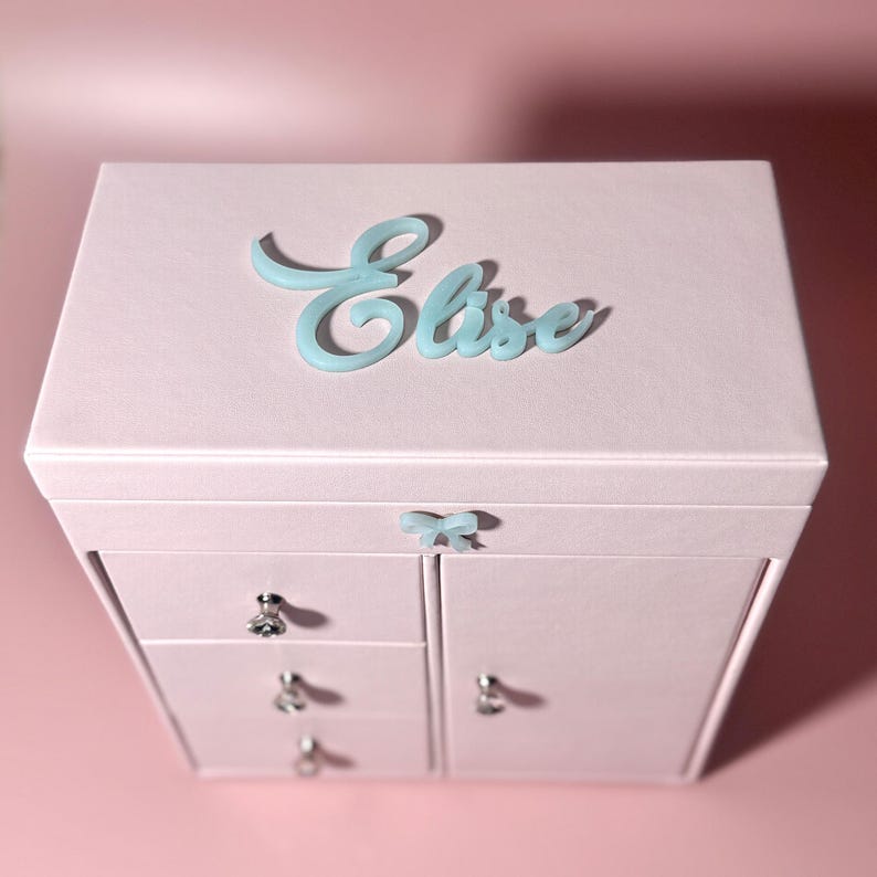 May include: A light pink jewelry box featuring a light blue script nameplate that reads Elise on the top lid. The box includes a small light blue bow accent on the front, three stacked drawers with silver knobs, and a side cabinet door.
