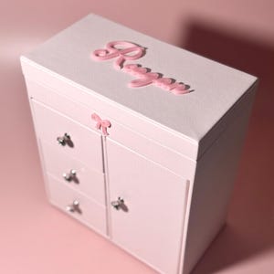 May include: A light pink jewelry box with a textured finish, featuring the name Regan in raised pink script on the top lid. The box includes three small drawers on the left side, a larger cabinet door on the right, and a small pink bow accent.