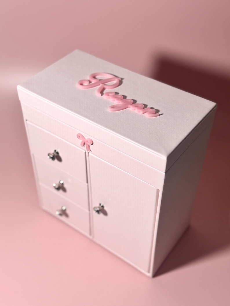May include: A light pink jewelry box with a textured finish, featuring the name Regan in raised pink script on the top lid. The box includes three small drawers on the left side, a larger cabinet door on the right, and a small pink bow accent.