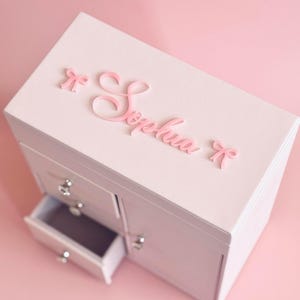 May include: A light pink jewelry box featuring the name Sophia in raised script lettering on the lid, flanked by two small bows. The text Style III SWEETHEART DOUBLE BOW custom name with 2 bows on the lid is visible at the top.