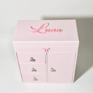 May include: A light pink jewelry box featuring a rectangular shape with a hinged lid. The name Luna is written in raised pink script on the top. The front includes three stacked drawers and a side door, each with a silver knob, and a small pink bow.