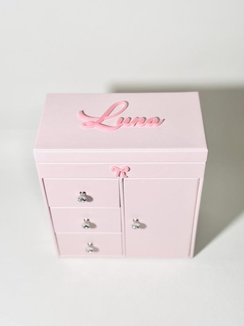 May include: A light pink jewelry box featuring a rectangular shape with a hinged lid. The name Luna is written in raised pink script on the top. The front includes three stacked drawers and a side door, each with a silver knob, and a small pink bow.