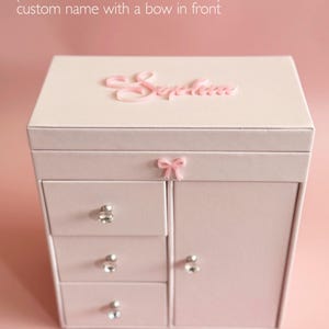 May include: A light pink jewelry box featuring the text Style II HEIRLOOM FRONT BOW (CUSTOMER FAVORITE!) custom name with a bow in front. The box has three drawers and a side cabinet, each with a clear crystal knob, and a pink bow accent.