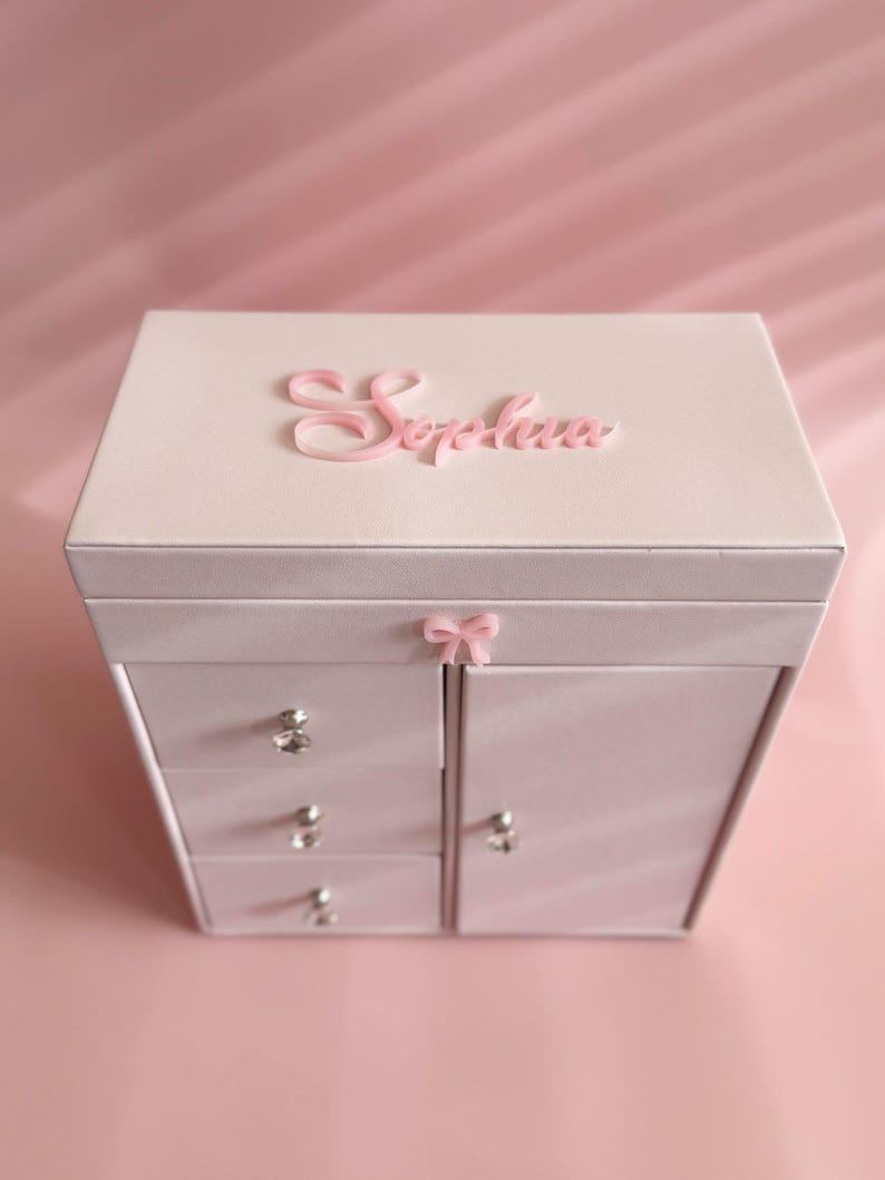 May include: A light pink jewelry box featuring the name Sophia in raised pink script on the top lid. The box includes three stacked drawers on the left and a single door on the right, each adorned with a small clear crystal knob and a pink bow.