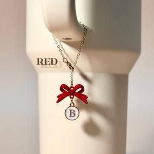 May include: A gold chain with a red bow and a white circle charm with the letter "B" in gold. The charm is hanging from a mug.