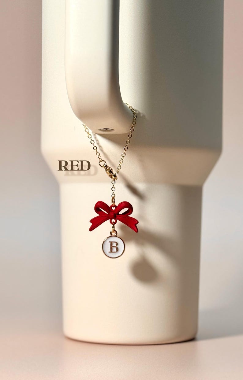 May include: A gold chain with a red bow and a white circle charm with the letter "B" in gold. The charm is hanging from a mug.