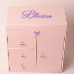 May include: A light pink jewelry box featuring a hinged lid with the name Blaire in purple script lettering. The front includes three stacked drawers on the left and a single door on the right, all adorned with clear, faceted crystal-style knobs.