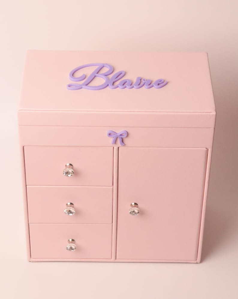 May include: A light pink jewelry box featuring a hinged lid with the name Blaire in purple script lettering. The front includes three stacked drawers on the left and a single door on the right, all adorned with clear, faceted crystal-style knobs.