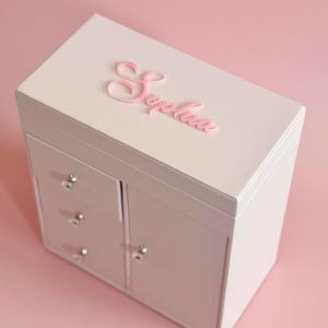 May include: A light pink jewelry box with a rectangular shape, featuring three small drawers and a side cabinet door with silver knobs. The top lid displays the name Sophia in raised pink script lettering. Text reads Style I SIGNATURE NAME.