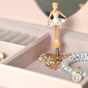 May include: A light pink jewelry box featuring a rotating ballerina figurine in a white tutu. Inside the velvet-lined compartment are gold-toned charms, including a starfish and a circular pendant with the letter D, and a blue floral bracelet.