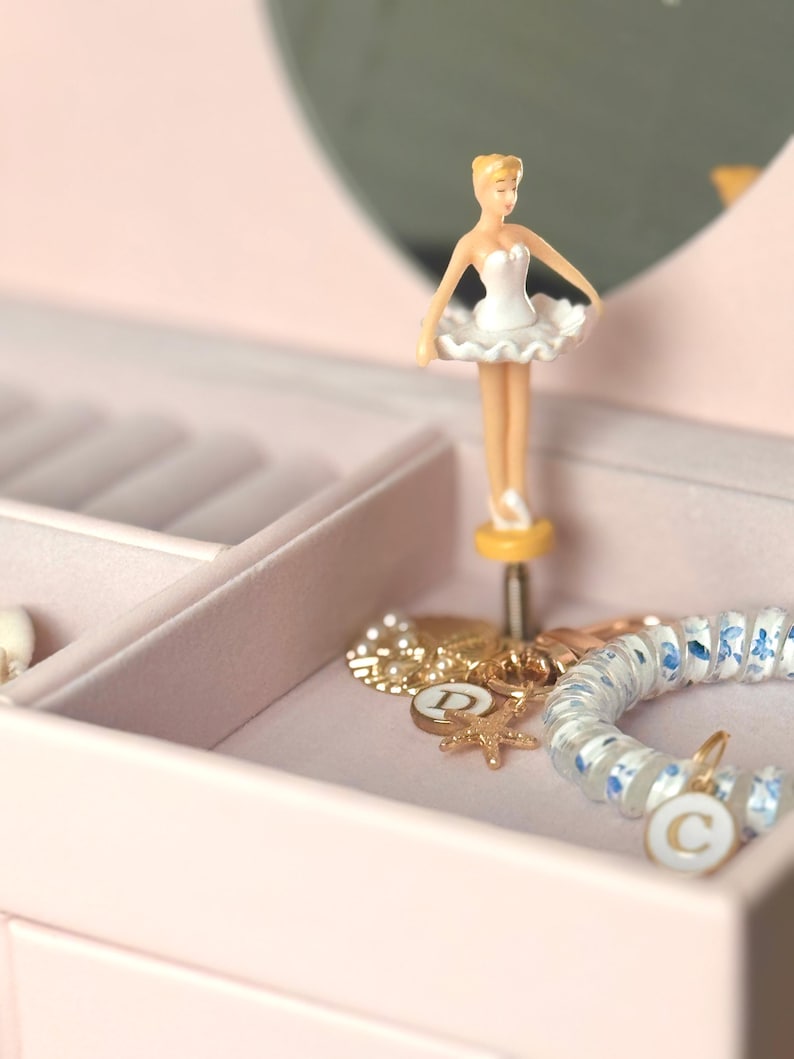 May include: A light pink jewelry box featuring a rotating ballerina figurine in a white tutu. Inside the velvet-lined compartment are gold-toned charms, including a starfish and a circular pendant with the letter D, and a blue floral bracelet.