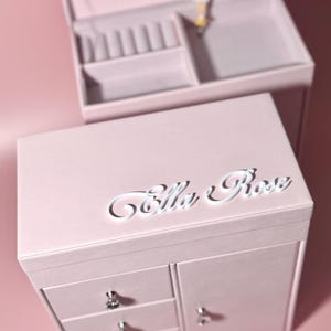 Puede incluir: Una caja de joyería rosa pálido con el nombre "Ella Rose" en escritura blanca en la tapa. La caja tiene cajones y una puerta de armario. La sección superior tiene un espejo y una figura de bailarina.