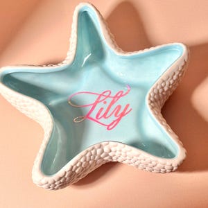 May include: A star-shaped ceramic trinket dish with a textured, bumpy white exterior and a smooth, light blue interior. The center of the dish features the name Lily written in a cursive, iridescent pink font that reflects light.