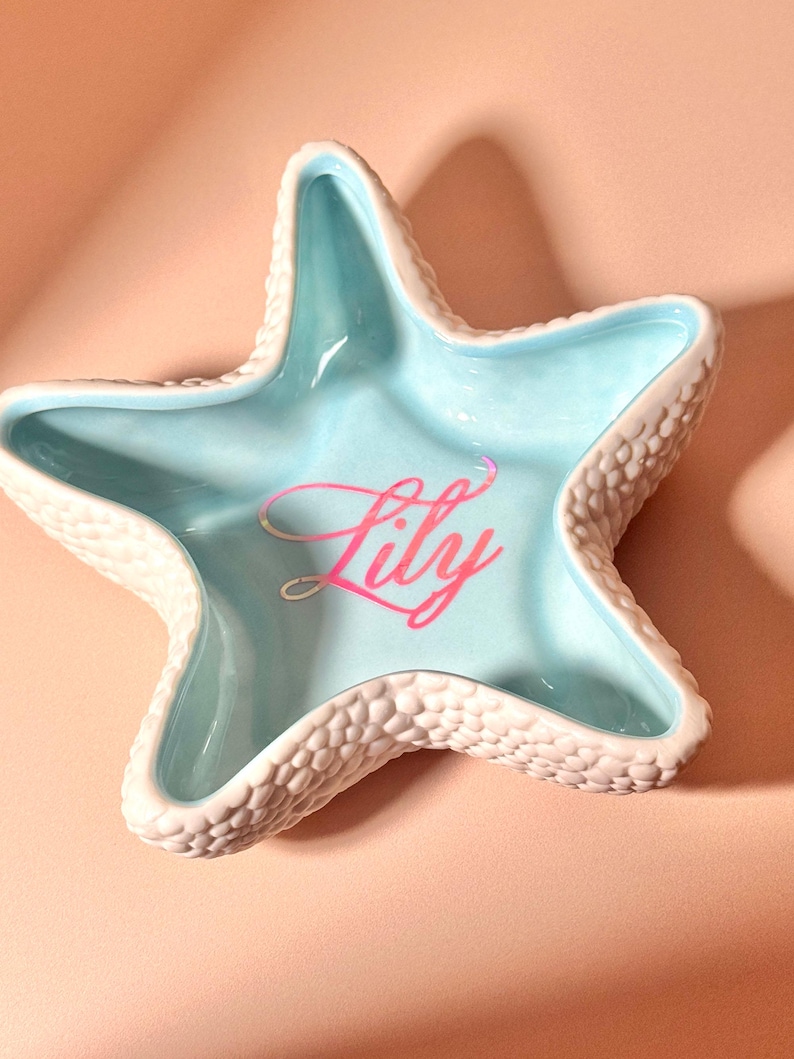 May include: A star-shaped ceramic trinket dish with a textured, bumpy white exterior and a smooth, light blue interior. The center of the dish features the name Lily written in a cursive, iridescent pink font that reflects light.