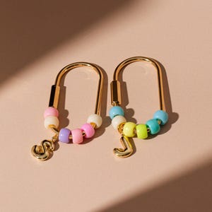 Peut inclure: Boucles d'oreilles dorées en forme de U. Chaque boucle d'oreille est ornée d'une rangée de perles colorées aux tons pastel et d'un charm en forme de lettre dorée. Sur fond rose clair.