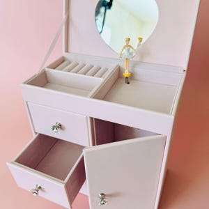 May include: A light pink jewelry box with a heart-shaped mirror on the inside lid. The interior features a ring holder, a small open compartment, and a rotating ballerina figurine. It includes two drawers and a side cabinet with crystal-style knobs.