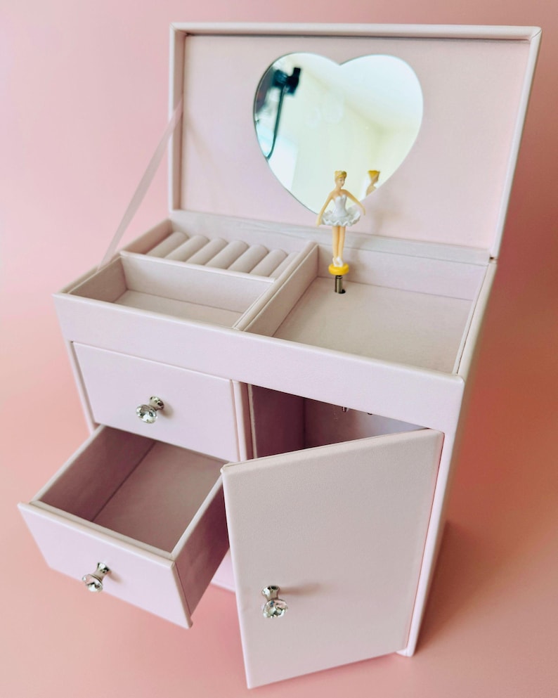 May include: A light pink jewelry box with a heart-shaped mirror on the inside lid. The interior features a ring holder, a small open compartment, and a rotating ballerina figurine. It includes two drawers and a side cabinet with crystal-style knobs.