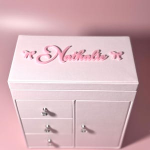 May include: A light pink jewelry box featuring the name Nathalie in raised pink script lettering on the top lid, flanked by two small pink bows. The box includes three stacked drawers on the left and a side door on the right, all with silver knobs.