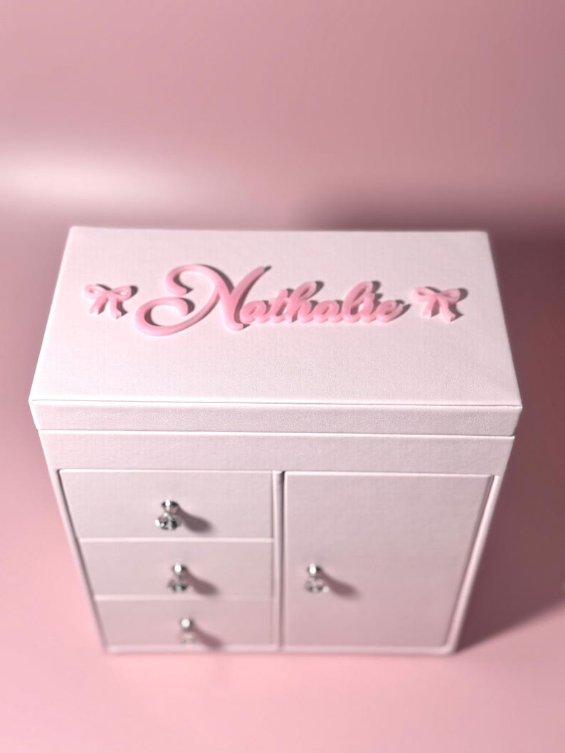 May include: A light pink jewelry box featuring the name Nathalie in raised pink script lettering on the top lid, flanked by two small pink bows. The box includes three stacked drawers on the left and a side door on the right, all with silver knobs.