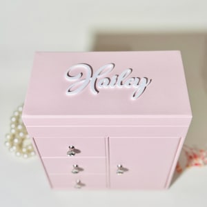 May include: A light pink jewelry box with a rectangular shape, featuring the name Hailey in white script lettering on the top lid. The front includes three small stacked drawers and one side door, each equipped with a clear crystal-like knob.