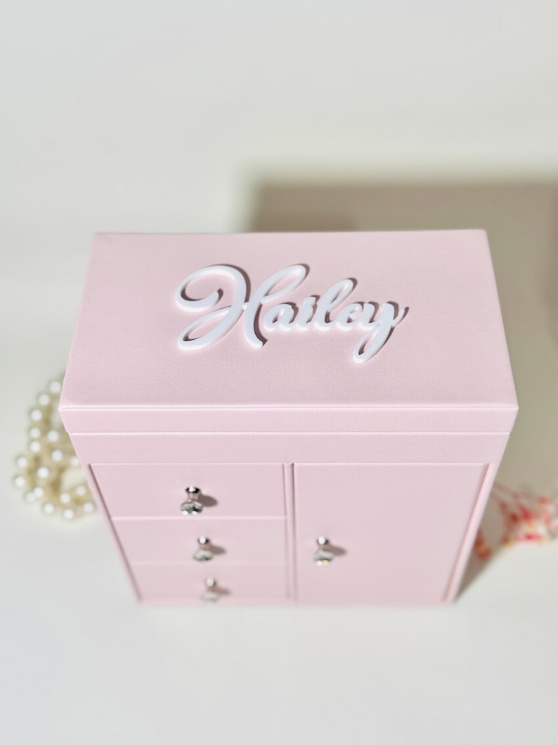 May include: A light pink jewelry box with a rectangular shape, featuring the name Hailey in white script lettering on the top lid. The front includes three small stacked drawers and one side door, each equipped with a clear crystal-like knob.