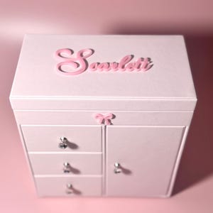 May include: A light pink jewelry box featuring the name Scarlett in raised pink lettering on the lid. The box includes three stacked drawers on the left and a side cabinet on the right, each adorned with clear crystal-style knobs and a small pink bow.