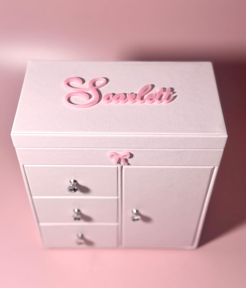 May include: A light pink jewelry box featuring the name Scarlett in raised pink lettering on the lid. The box includes three stacked drawers on the left and a side cabinet on the right, each adorned with clear crystal-style knobs and a small pink bow.