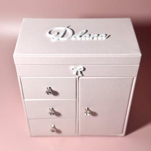 May include: A light pink jewelry box featuring a white script name Delana on the top lid, a small white bow accent on the front, three drawers with clear crystal-like knobs, and a side cabinet door with a matching crystal-like knob.